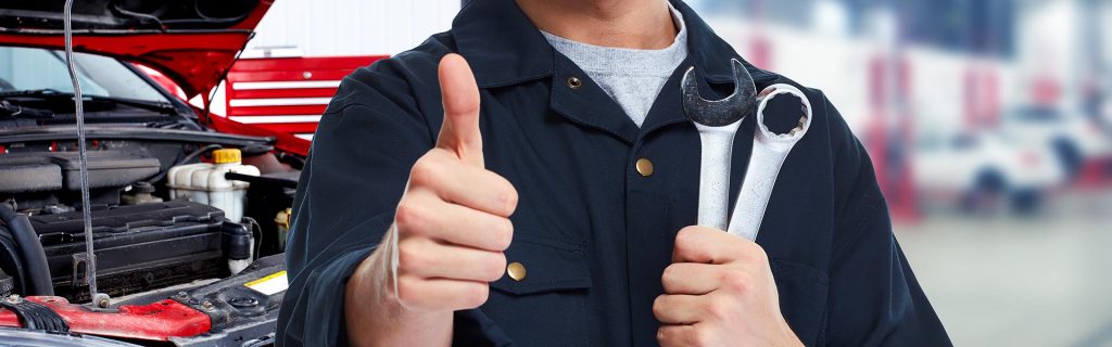 Mechanic holding wrenches and giving a thumbs-up in front of an open car hood.